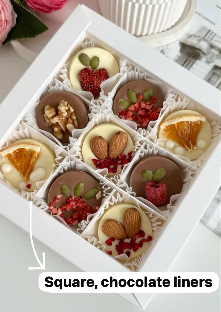 A boxed assortment of decorated chocolate squares and chocolate liners arranged in a white gift box, with pink flowers and a white cupcake in the background. Text on the image reads 'Square, chocolate liners'.