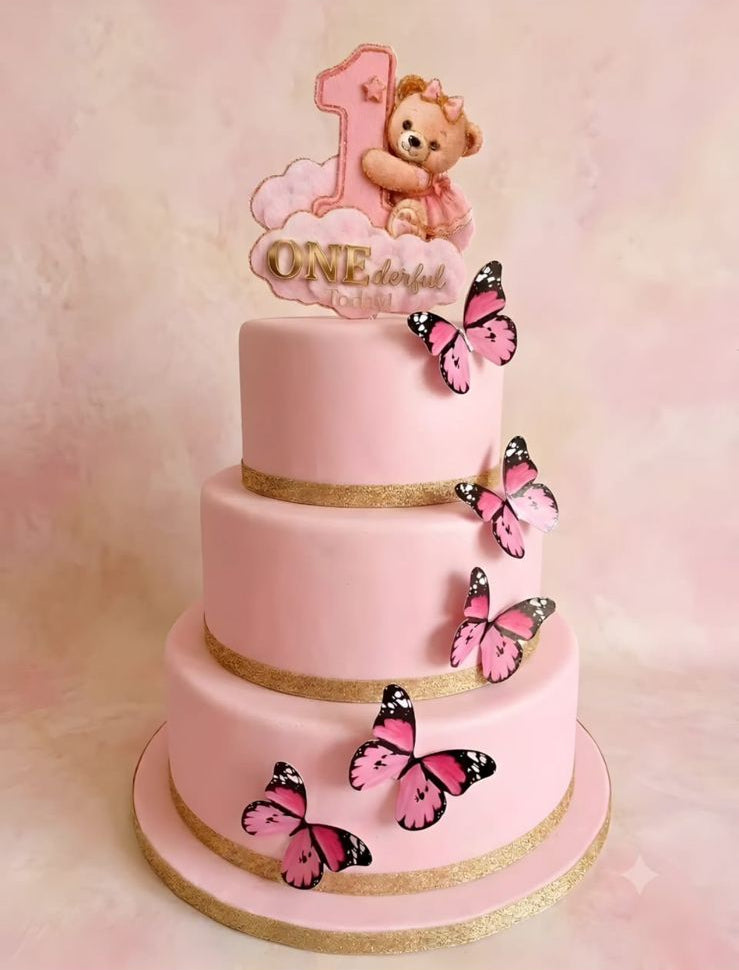 A three-tier pink cake decorated with gold bands, pink and black patterned butterflies, and a teddy bear 'ONE' topper, shown against a soft pink background.